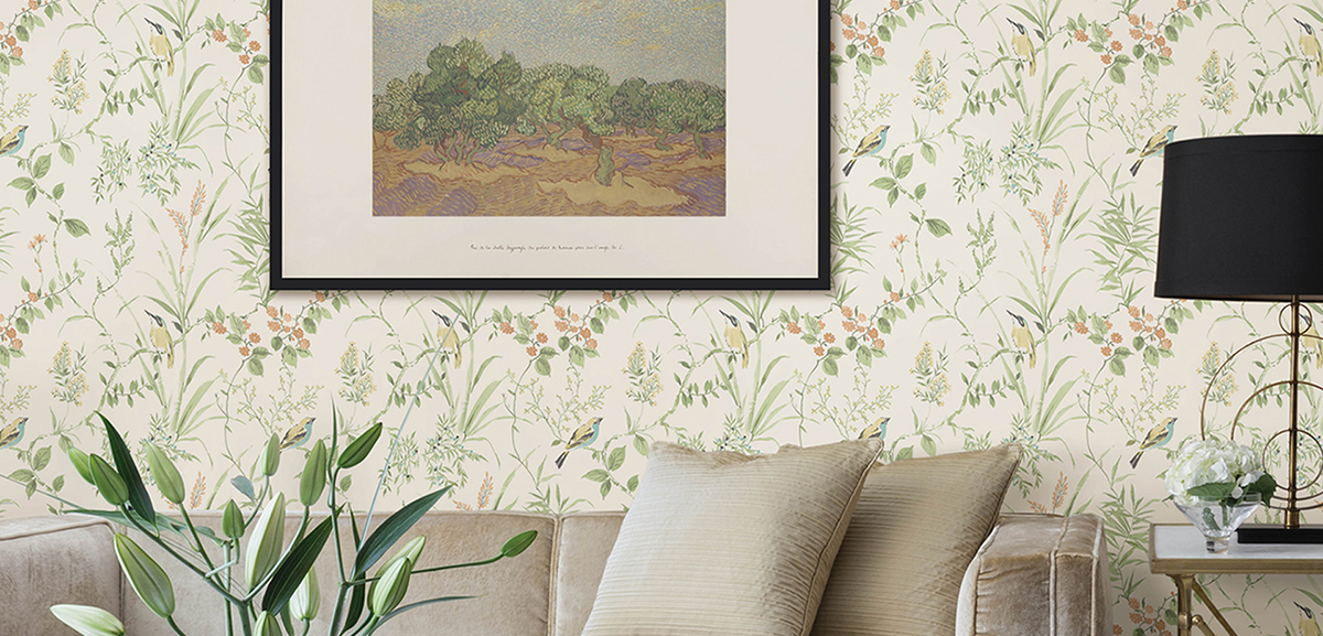 A beige sofa with striped pillows sits against a wall with floral wallpaper. Above the sofa hangs a framed painting of trees. A black table lamp and a vase of green plants are on a side table beside the sofa.