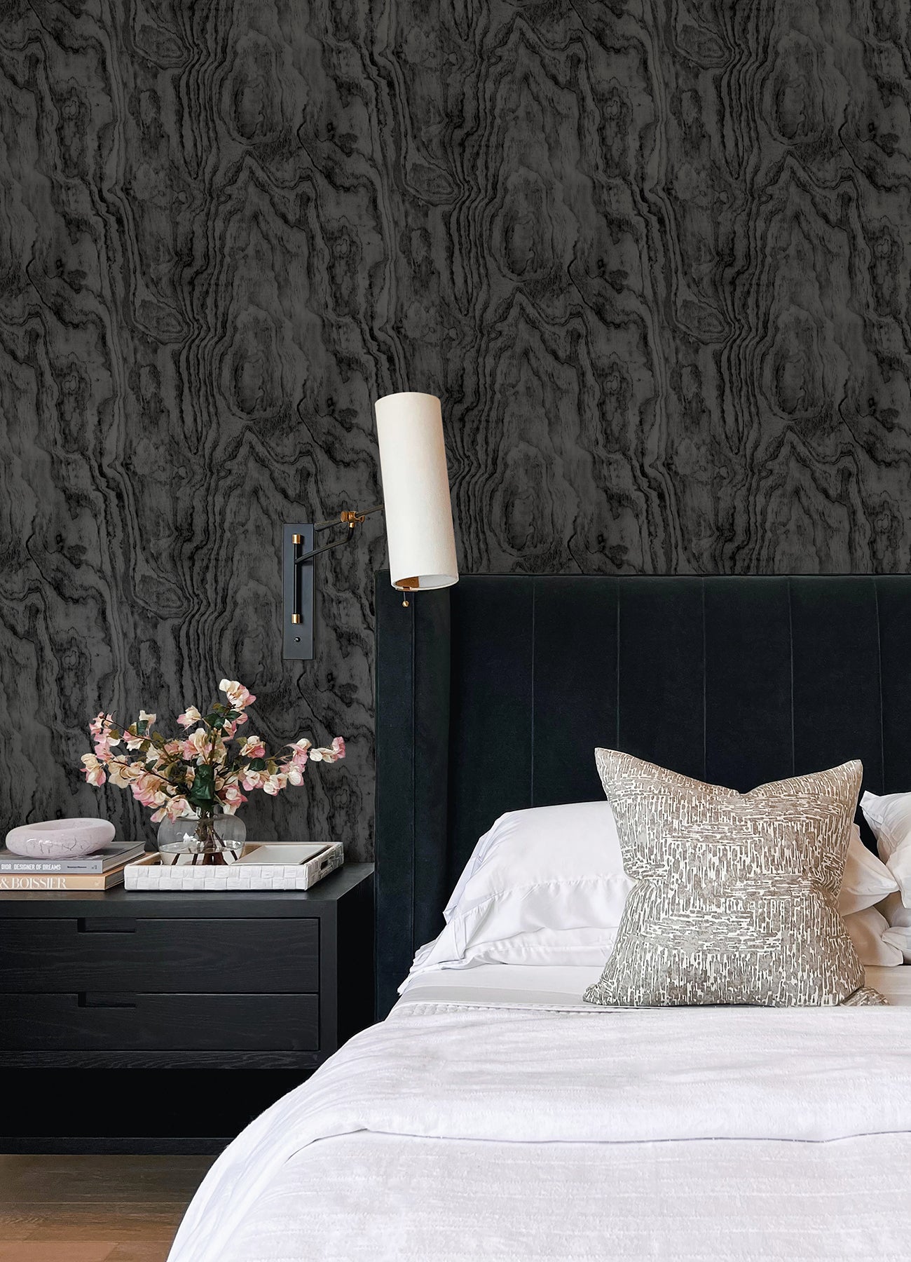 A modern bedroom features a dark, textured accent wall created with Ashley Stark Home Sloane Wood Peel and Stick Wallpaper by WallPops. A black upholstered bed with white bedding and a patterned pillow is complemented by a black nightstand that holds books, a towel, and a vase of pink flowers, all situated beneath a sleek wall-mounted lamp.