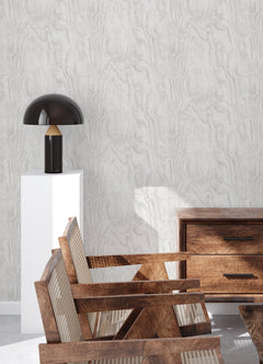 A modern bedroom features a dark, textured accent wall created with Ashley Stark Home Sloane Wood Peel and Stick Wallpaper by WallPops. A black upholstered bed with white bedding and a patterned pillow is complemented by a black nightstand that holds books, a towel, and a vase of pink flowers, all situated beneath a sleek wall-mounted lamp.