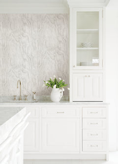 A modern bedroom features a dark, textured accent wall created with Ashley Stark Home Sloane Wood Peel and Stick Wallpaper by WallPops. A black upholstered bed with white bedding and a patterned pillow is complemented by a black nightstand that holds books, a towel, and a vase of pink flowers, all situated beneath a sleek wall-mounted lamp.