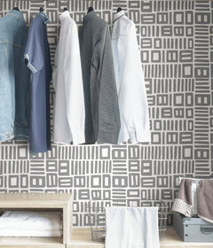 Beneath dark gray cabinets, a backsplash is enhanced with WallPops Black Pepper Paperie Traverse Peel and Stick Wallpaper, showcasing a bold geometric gray and white square pattern reminiscent of striking wallpaper. On the white countertop sit three white canisters with wooden lids alongside a large white bowl filled with green apples.