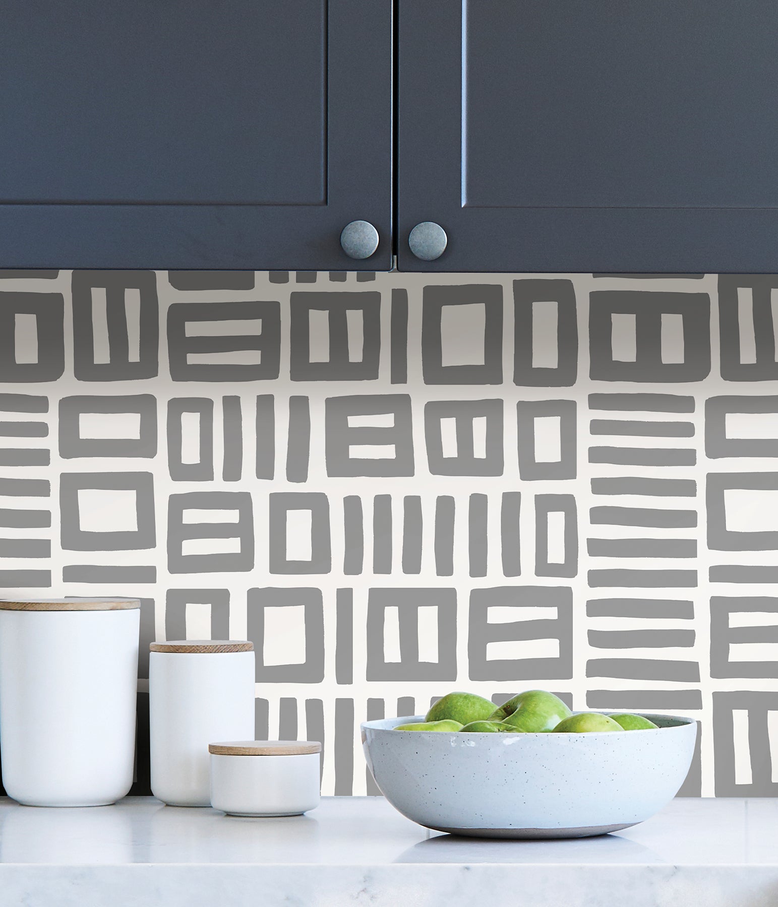 Beneath dark gray cabinets, a backsplash is enhanced with WallPops Black Pepper Paperie Traverse Peel and Stick Wallpaper, showcasing a bold geometric gray and white square pattern reminiscent of striking wallpaper. On the white countertop sit three white canisters with wooden lids alongside a large white bowl filled with green apples.