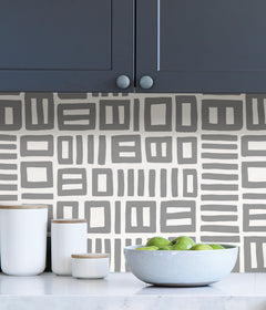 Beneath dark gray cabinets, a backsplash is enhanced with WallPops Black Pepper Paperie Traverse Peel and Stick Wallpaper, showcasing a bold geometric gray and white square pattern reminiscent of striking wallpaper. On the white countertop sit three white canisters with wooden lids alongside a large white bowl filled with green apples.