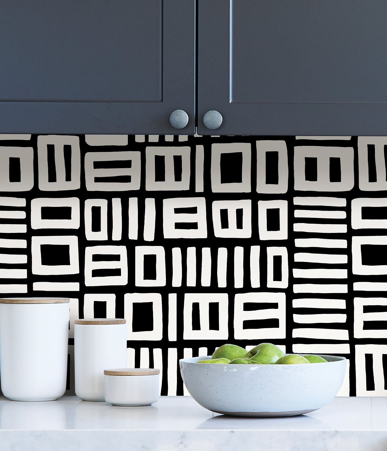 Beneath dark gray cabinets, a backsplash is enhanced with WallPops Black Pepper Paperie Traverse Peel and Stick Wallpaper, showcasing a bold geometric gray and white square pattern reminiscent of striking wallpaper. On the white countertop sit three white canisters with wooden lids alongside a large white bowl filled with green apples.
