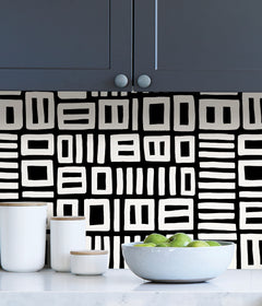 Beneath dark gray cabinets, a backsplash is enhanced with WallPops Black Pepper Paperie Traverse Peel and Stick Wallpaper, showcasing a bold geometric gray and white square pattern reminiscent of striking wallpaper. On the white countertop sit three white canisters with wooden lids alongside a large white bowl filled with green apples.