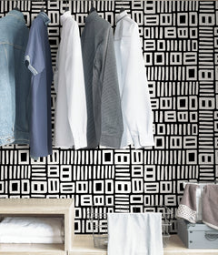 Beneath dark gray cabinets, a backsplash is enhanced with WallPops Black Pepper Paperie Traverse Peel and Stick Wallpaper, showcasing a bold geometric gray and white square pattern reminiscent of striking wallpaper. On the white countertop sit three white canisters with wooden lids alongside a large white bowl filled with green apples.