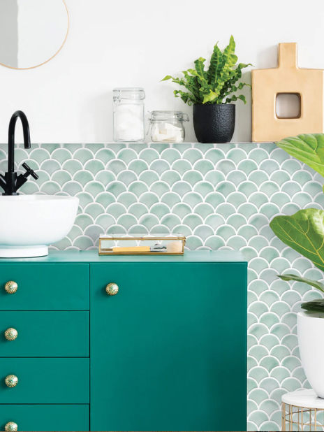 Modern bathroom scene with teal cabinet, white vessel sink, black faucet, and brass knobs. Green fish-scale tile backsplash, round mirror, plants, jars, a wooden tray, and decor accents create a stylish, fresh look. A fiddle leaf fig is in the bottom right corner.