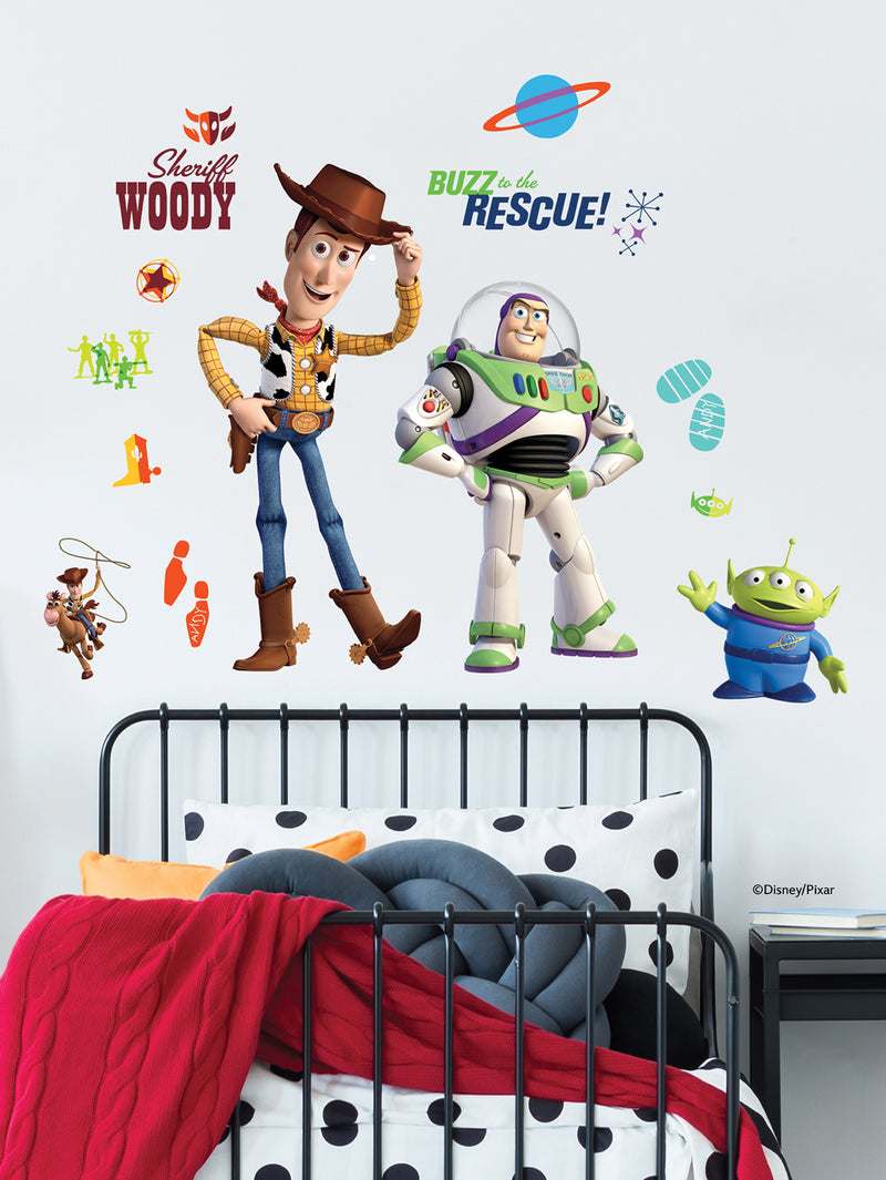 A children’s bedroom wall decorated with large stickers of Woody, Buzz Lightyear, and an alien from Toy Story. The black metal bed frame has black and white polka dot bedding and a red blanket. The wall also has colorful Toy Story-themed graphics and phrases.