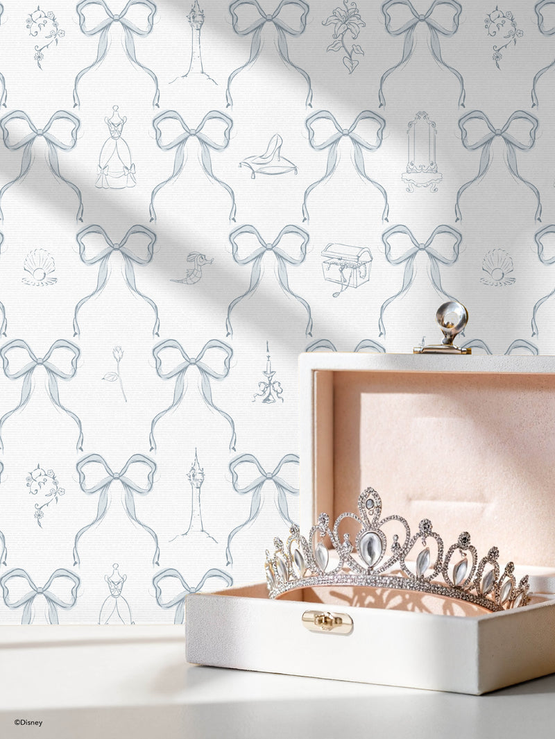 A jeweled tiara sits in an open, cream-colored velvet box. The background wallpaper is patterned with delicate, light blue bows and Disney princess-themed sketches, including a slipper, rose, and candelabra. Light casts soft shadows across the scene.