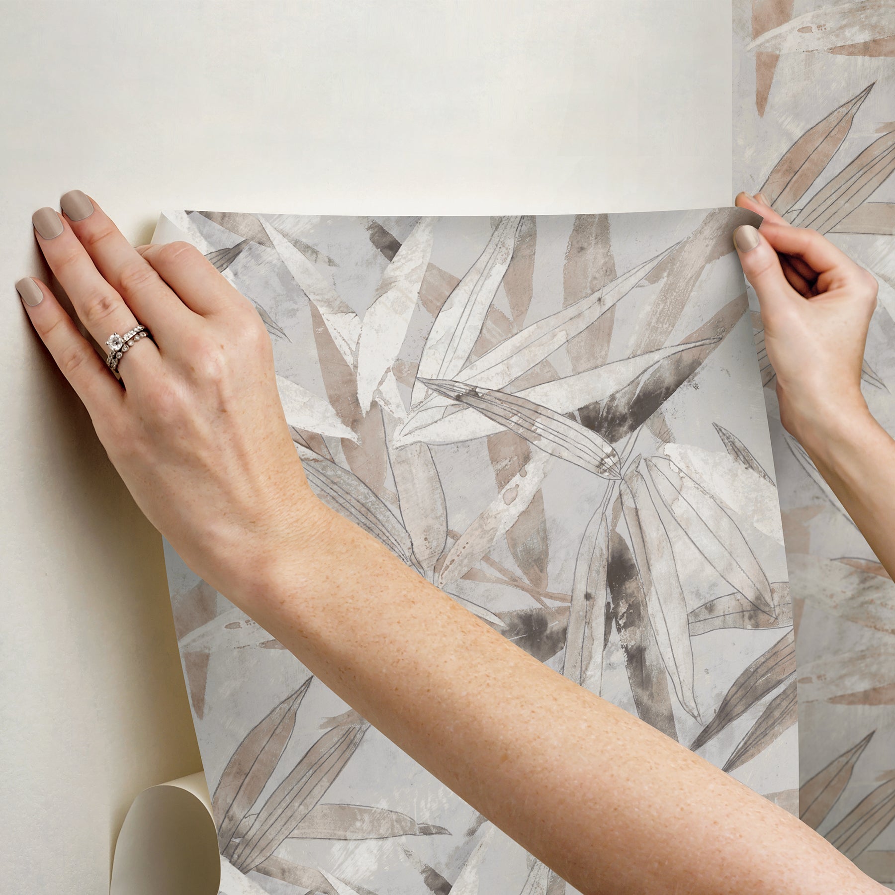 A person peels back a corner of the WallPops Egypt Sherrod Bali Bamboo Peel and Stick Wallpaper, which features abstract leaf designs in beige, gray, and blush tones. The wallpaper is displayed on a wooden surface, and the person wears a wedding ring and another ring on their left hand’s ring finger.