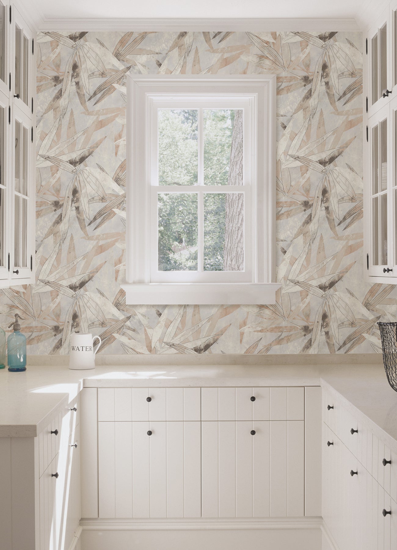 A person peels back a corner of the WallPops Egypt Sherrod Bali Bamboo Peel and Stick Wallpaper, which features abstract leaf designs in beige, gray, and blush tones. The wallpaper is displayed on a wooden surface, and the person wears a wedding ring and another ring on their left hand’s ring finger.