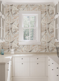 A person peels back a corner of the WallPops Egypt Sherrod Bali Bamboo Peel and Stick Wallpaper, which features abstract leaf designs in beige, gray, and blush tones. The wallpaper is displayed on a wooden surface, and the person wears a wedding ring and another ring on their left hand’s ring finger.