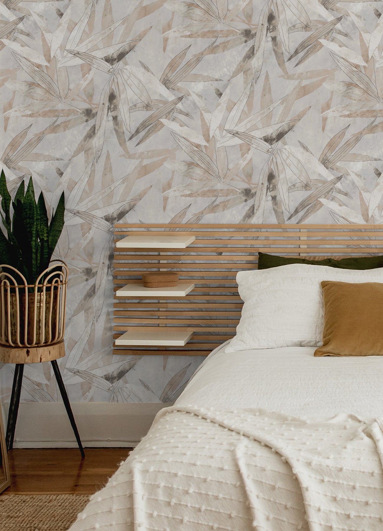 A person peels back a corner of the WallPops Egypt Sherrod Bali Bamboo Peel and Stick Wallpaper, which features abstract leaf designs in beige, gray, and blush tones. The wallpaper is displayed on a wooden surface, and the person wears a wedding ring and another ring on their left hand’s ring finger.