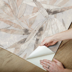 A person peels back a corner of the WallPops Egypt Sherrod Bali Bamboo Peel and Stick Wallpaper, which features abstract leaf designs in beige, gray, and blush tones. The wallpaper is displayed on a wooden surface, and the person wears a wedding ring and another ring on their left hand’s ring finger.