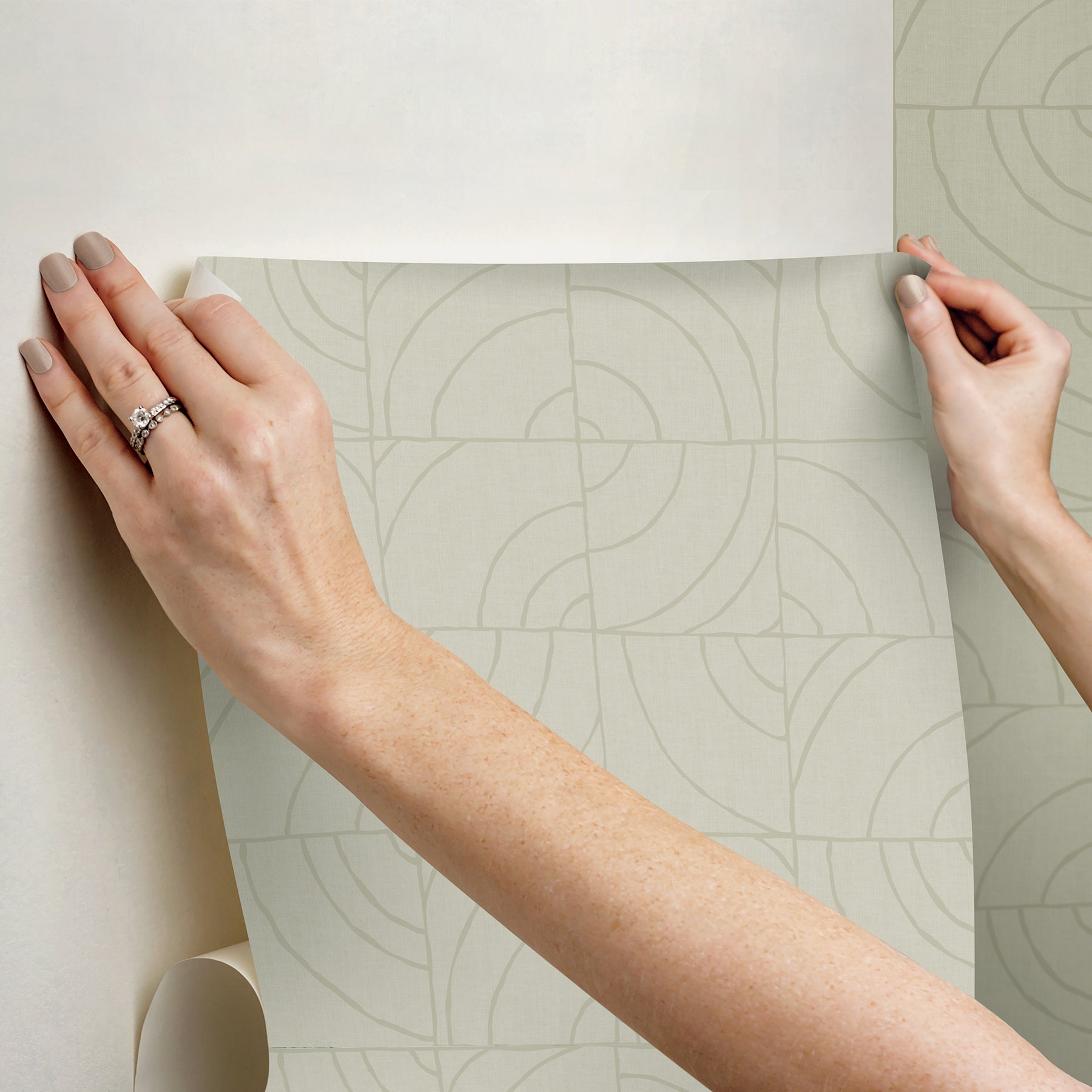 A person’s hands peel back a corner of WallPops Egypt Sherrod Batik Blok Peel and Stick Wallpaper, featuring abstract curved lines on a beige background, revealing its adhesive backing. The wallpaper rests on a wooden surface, and the person wears a ring on their left hand.