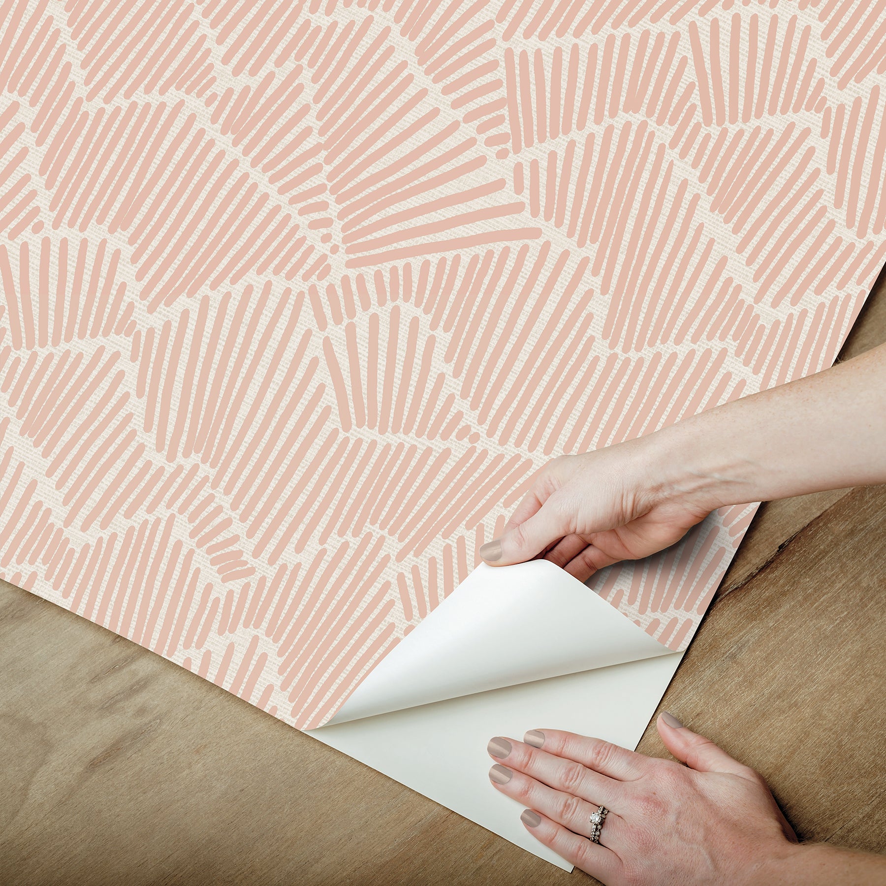 Two hands peel back a sheet of WallPops Egypt Sherrod Ridge & Valley Peel and Stick Wallpaper, which features a white abstract geometric line pattern on a beige background. The wallpaper is partially lifted from a wooden surface, and the person’s left hand is adorned with a ring.