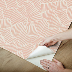 Two hands peel back a sheet of WallPops Egypt Sherrod Ridge & Valley Peel and Stick Wallpaper, which features a white abstract geometric line pattern on a beige background. The wallpaper is partially lifted from a wooden surface, and the person’s left hand is adorned with a ring.