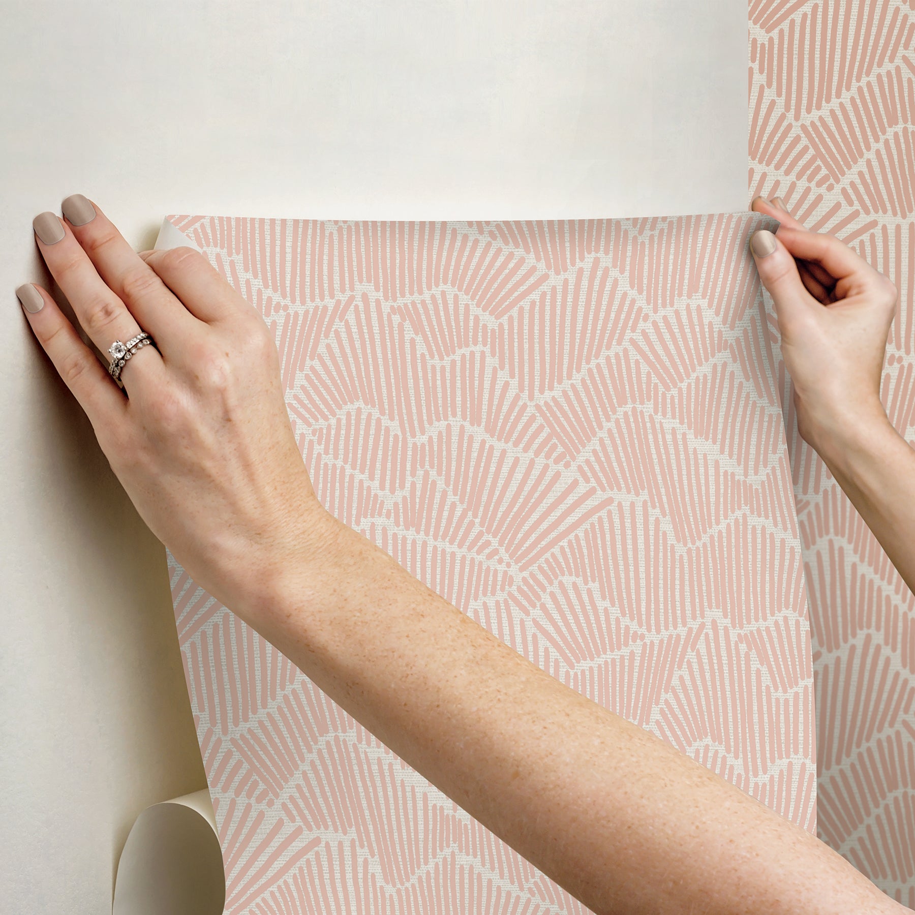 Two hands peel back a sheet of WallPops Egypt Sherrod Ridge & Valley Peel and Stick Wallpaper, which features a white abstract geometric line pattern on a beige background. The wallpaper is partially lifted from a wooden surface, and the person’s left hand is adorned with a ring.