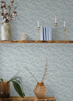 Two hands peel back a sheet of WallPops Egypt Sherrod Ridge & Valley Peel and Stick Wallpaper, which features a white abstract geometric line pattern on a beige background. The wallpaper is partially lifted from a wooden surface, and the person’s left hand is adorned with a ring.