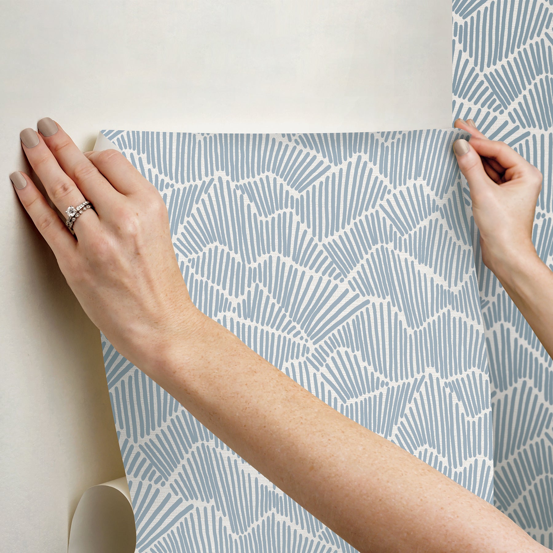 Two hands peel back a sheet of WallPops Egypt Sherrod Ridge & Valley Peel and Stick Wallpaper, which features a white abstract geometric line pattern on a beige background. The wallpaper is partially lifted from a wooden surface, and the person’s left hand is adorned with a ring.