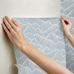 Two hands peel back a sheet of WallPops Egypt Sherrod Ridge & Valley Peel and Stick Wallpaper, which features a white abstract geometric line pattern on a beige background. The wallpaper is partially lifted from a wooden surface, and the person’s left hand is adorned with a ring.