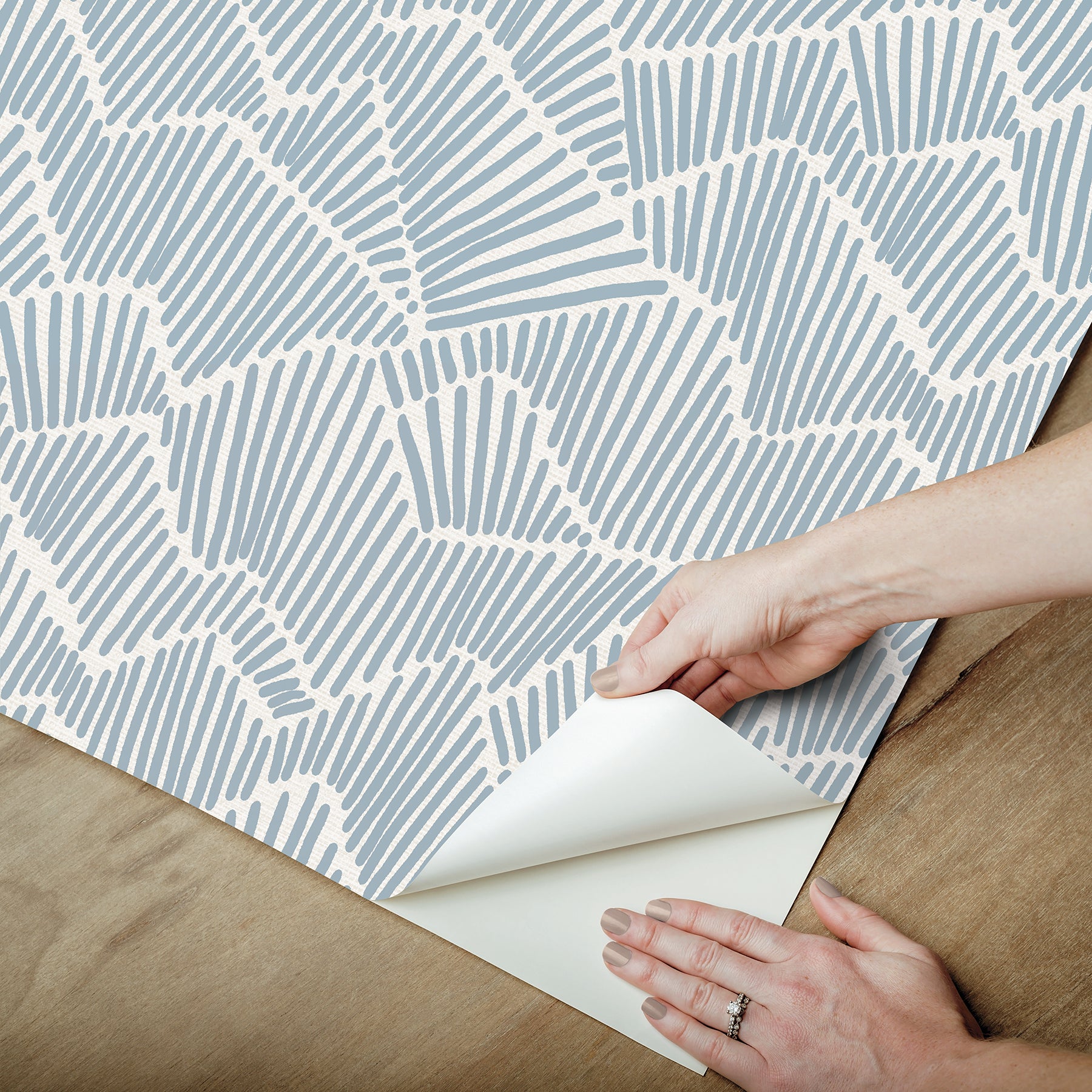 Two hands peel back a sheet of WallPops Egypt Sherrod Ridge & Valley Peel and Stick Wallpaper, which features a white abstract geometric line pattern on a beige background. The wallpaper is partially lifted from a wooden surface, and the person’s left hand is adorned with a ring.
