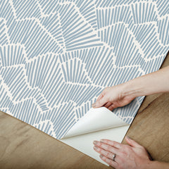 Two hands peel back a sheet of WallPops Egypt Sherrod Ridge & Valley Peel and Stick Wallpaper, which features a white abstract geometric line pattern on a beige background. The wallpaper is partially lifted from a wooden surface, and the person’s left hand is adorned with a ring.