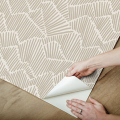 Two hands peel back a sheet of WallPops Egypt Sherrod Ridge & Valley Peel and Stick Wallpaper, which features a white abstract geometric line pattern on a beige background. The wallpaper is partially lifted from a wooden surface, and the person’s left hand is adorned with a ring.