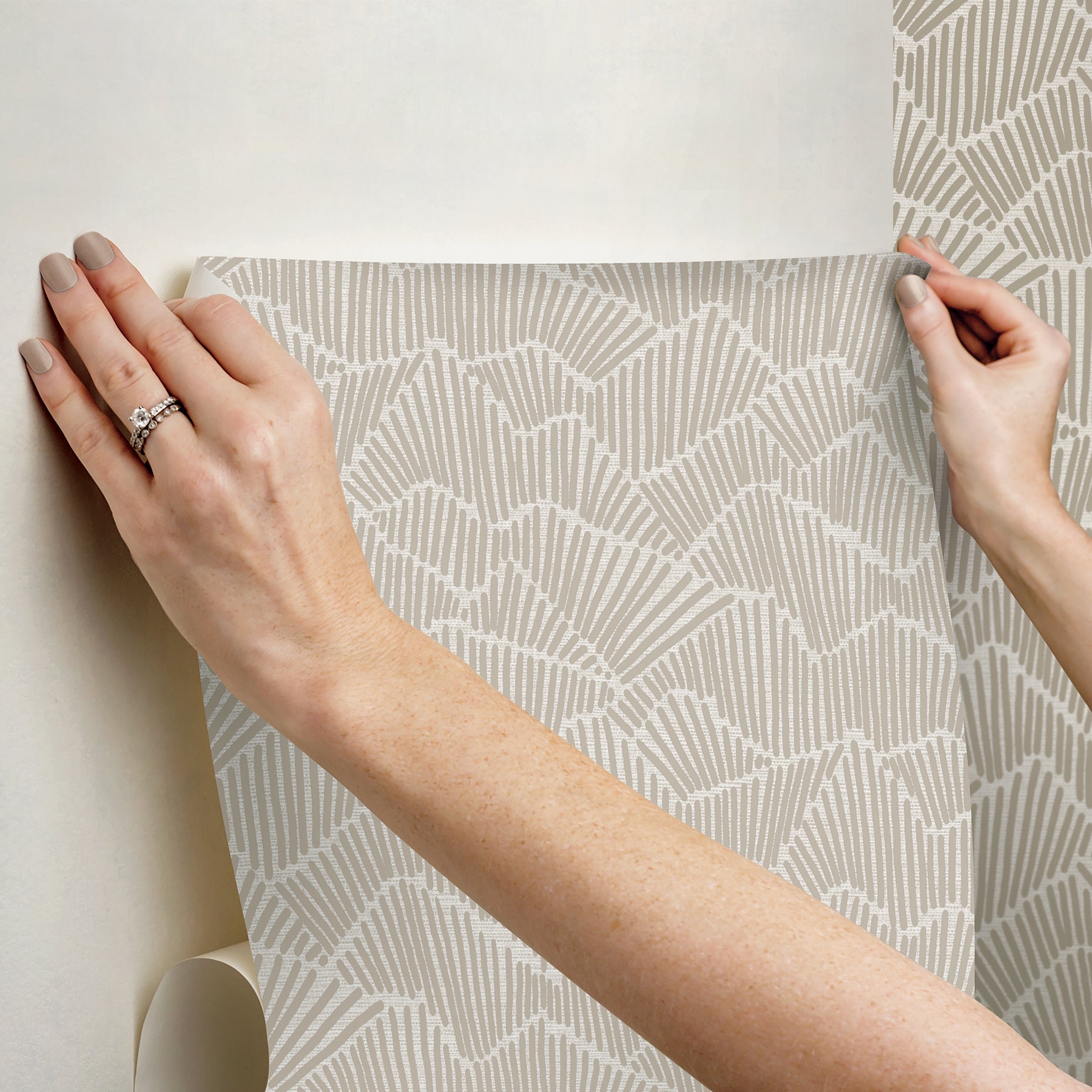 Two hands peel back a sheet of WallPops Egypt Sherrod Ridge & Valley Peel and Stick Wallpaper, which features a white abstract geometric line pattern on a beige background. The wallpaper is partially lifted from a wooden surface, and the person’s left hand is adorned with a ring.