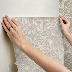 Two hands peel back a sheet of WallPops Egypt Sherrod Ridge & Valley Peel and Stick Wallpaper, which features a white abstract geometric line pattern on a beige background. The wallpaper is partially lifted from a wooden surface, and the person’s left hand is adorned with a ring.