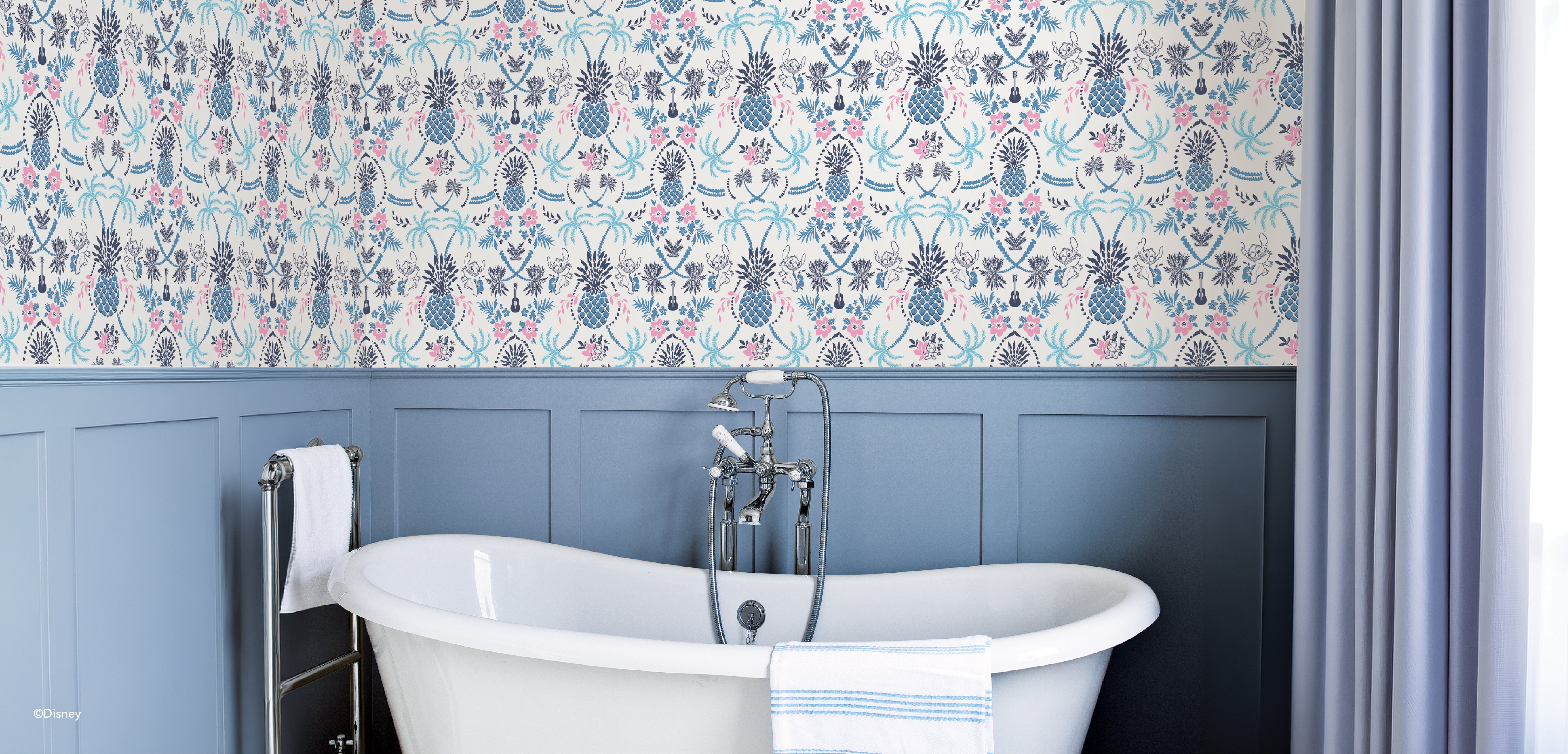 A white freestanding bathtub with a towel draped over the side sits against blue wainscoting and wallpaper featuring blue, pink, and grey floral and pineapple patterns. Chrome faucet and shower fixtures are included; light streams through a curtained window.