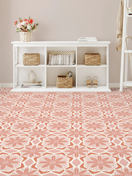 White cubby shelf with baskets, books, a striped pillow, and shoes sits against a beige wall. A vase with pink flowers is on top. The floor has a bold red and white geometric floral pattern. A white ladder shelf is partially visible on the right.