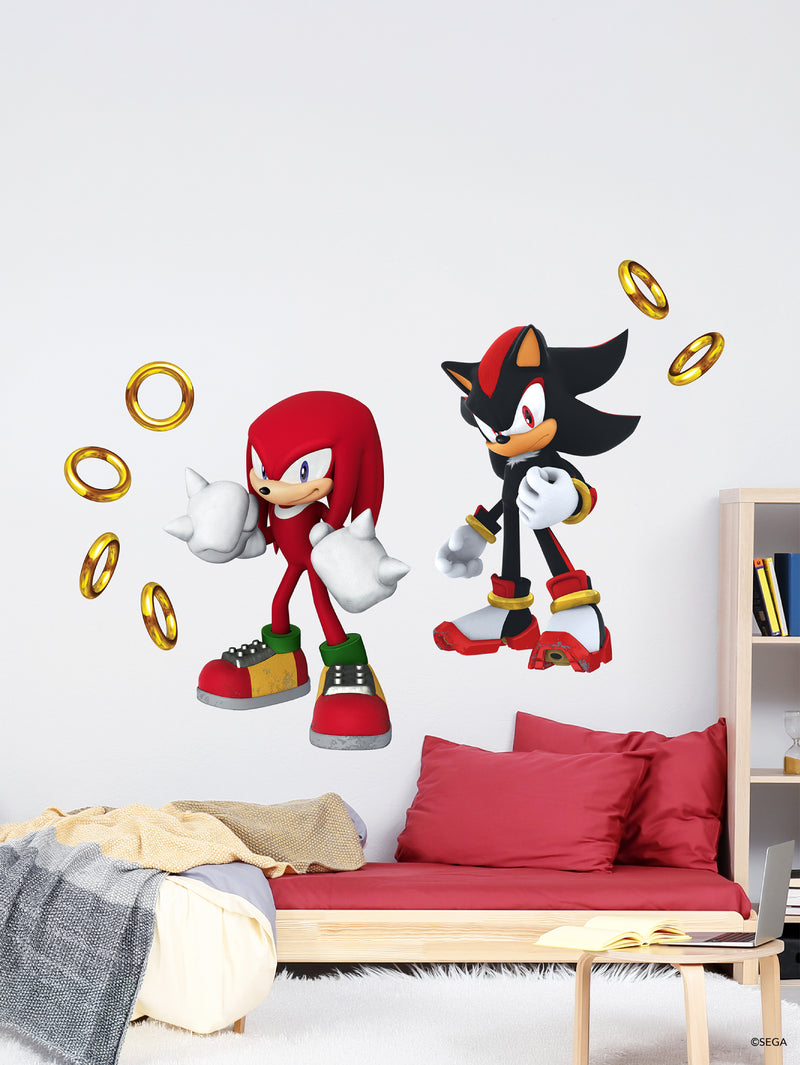 Wall decals of Sonic characters Knuckles and Shadow stand above a red bed with pillows. Gold rings float around them. A beige blanket lies on the bed, and a wooden shelf with books is on the right, emphasizing a playful, themed bedroom.