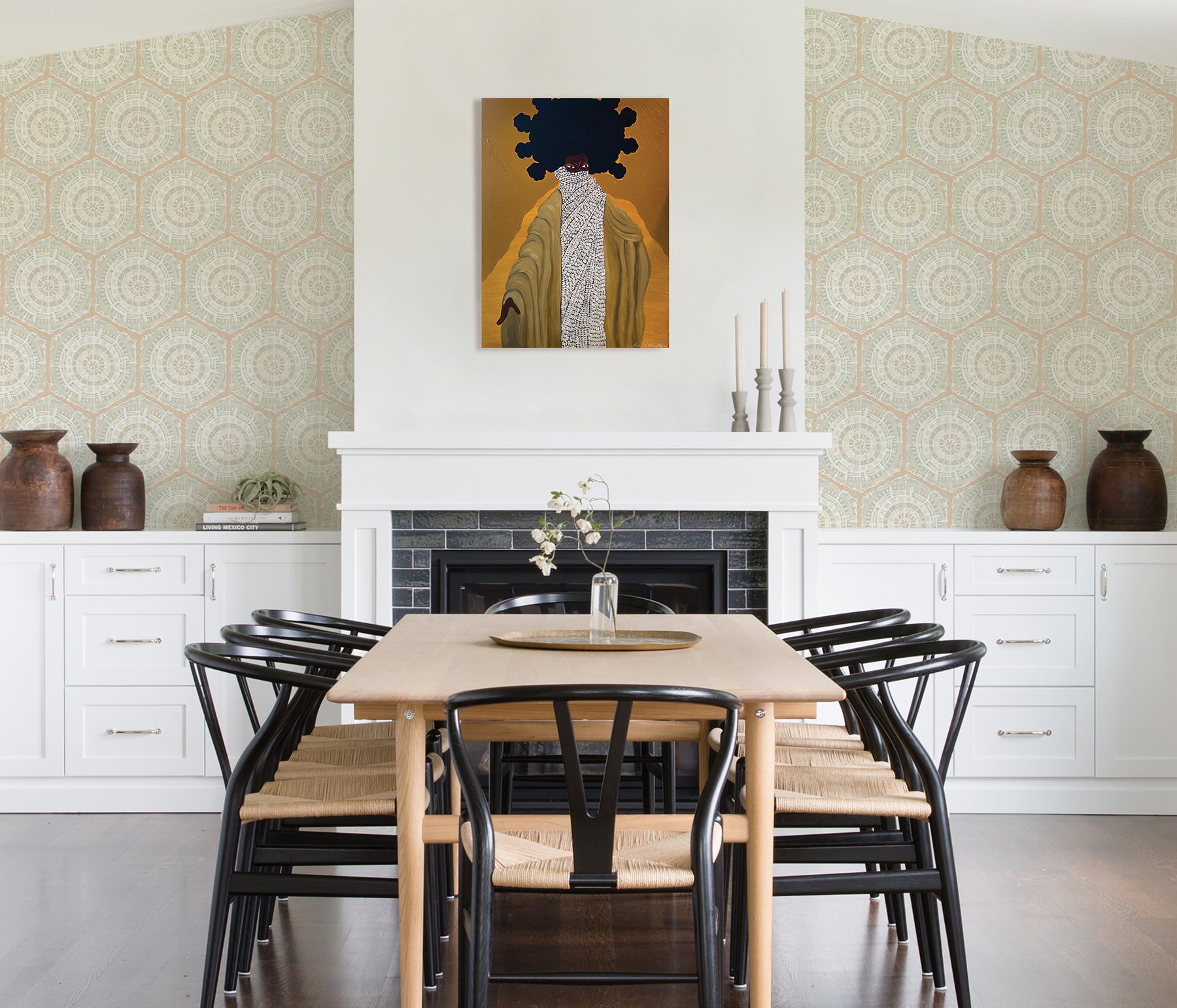 A modern dining room featuring a wooden table, eight black chairs, white built-in cabinets, and blue LiLi WhittWhitt Harmony Peel and Stick Wallpaper by WallPops. Above a white fireplace hangs a colorful abstract portrait. The space is accented with decorative pottery, candles, and a single-stem vase.