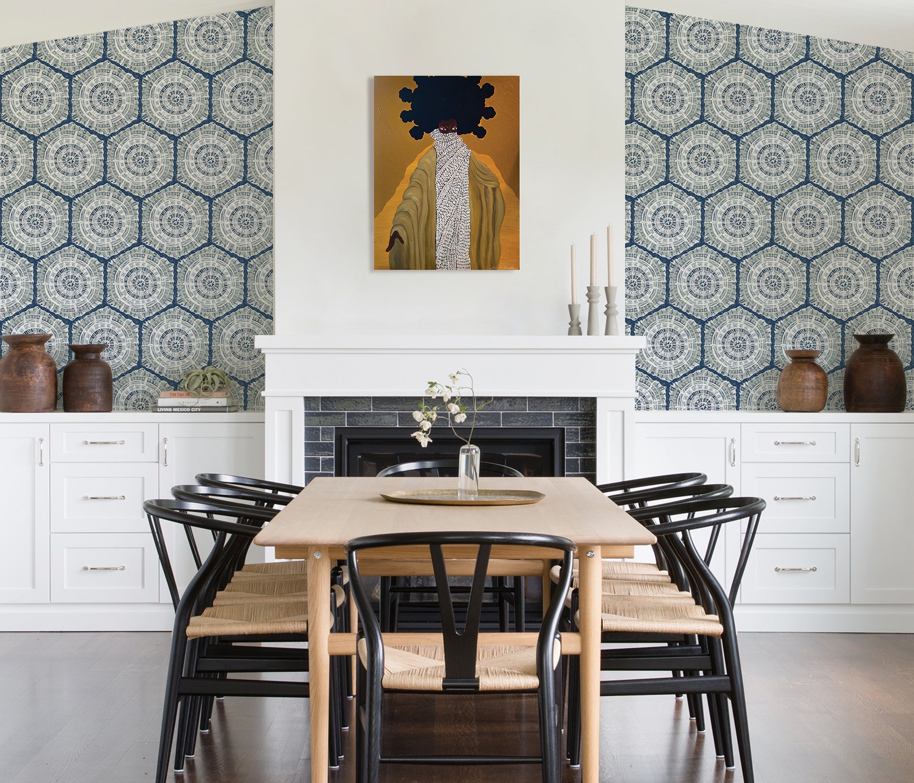 A modern dining room featuring a wooden table, eight black chairs, white built-in cabinets, and blue LiLi WhittWhitt Harmony Peel and Stick Wallpaper by WallPops. Above a white fireplace hangs a colorful abstract portrait. The space is accented with decorative pottery, candles, and a single-stem vase.