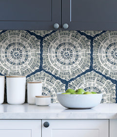 A modern dining room featuring a wooden table, eight black chairs, white built-in cabinets, and blue LiLi WhittWhitt Harmony Peel and Stick Wallpaper by WallPops. Above a white fireplace hangs a colorful abstract portrait. The space is accented with decorative pottery, candles, and a single-stem vase.