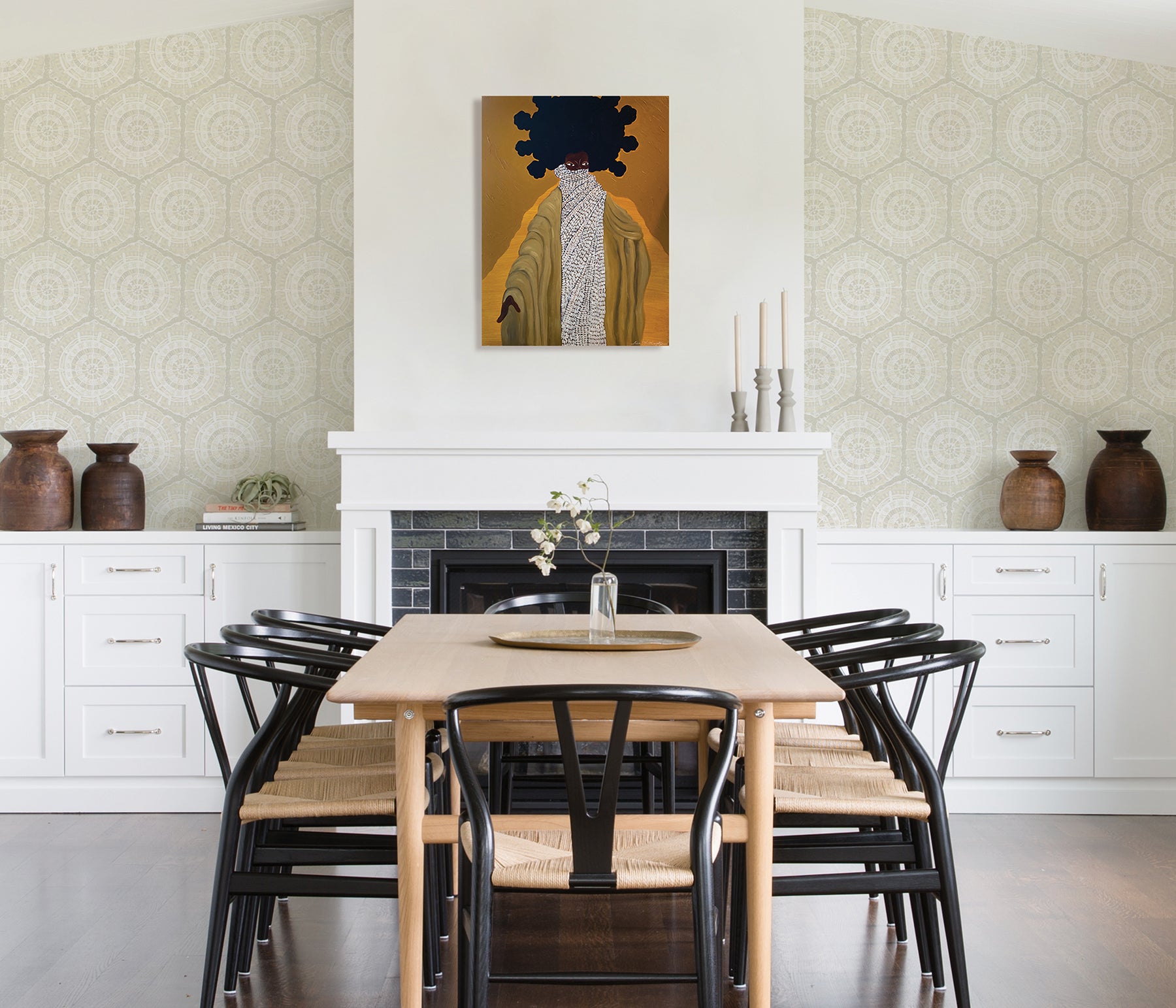 A modern dining room featuring a wooden table, eight black chairs, white built-in cabinets, and blue LiLi WhittWhitt Harmony Peel and Stick Wallpaper by WallPops. Above a white fireplace hangs a colorful abstract portrait. The space is accented with decorative pottery, candles, and a single-stem vase.