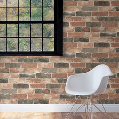 A modern white chair sits on a wooden floor against a Newport Reclaimed Brick Peel And Stick Wallpaper accent wall, with a large window revealing lush green trees outside.