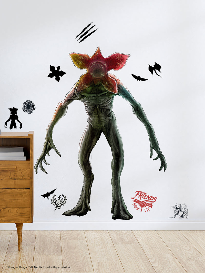 A large wall decal of a Demogorgon from Stranger Things stands menacingly on a white wall, surrounded by black monster silhouettes, claw marks, and the “Friends Don’t Lie” phrase in red. Part of a wooden dresser is visible on the left.