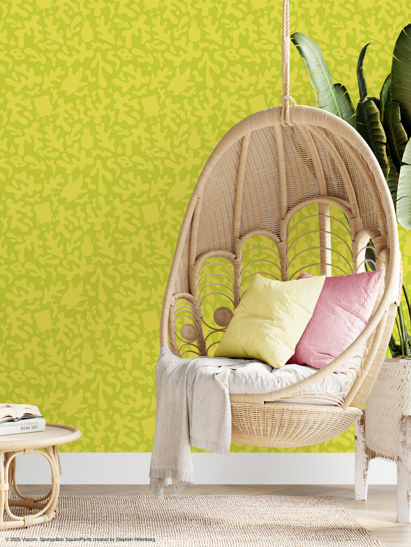 A wicker hanging chair with pastel pillows and a light throw sits next to a green leafy plant. The yellow-green wall behind features an abstract leaf pattern, and a small table with a glass top is partly visible in the foreground.