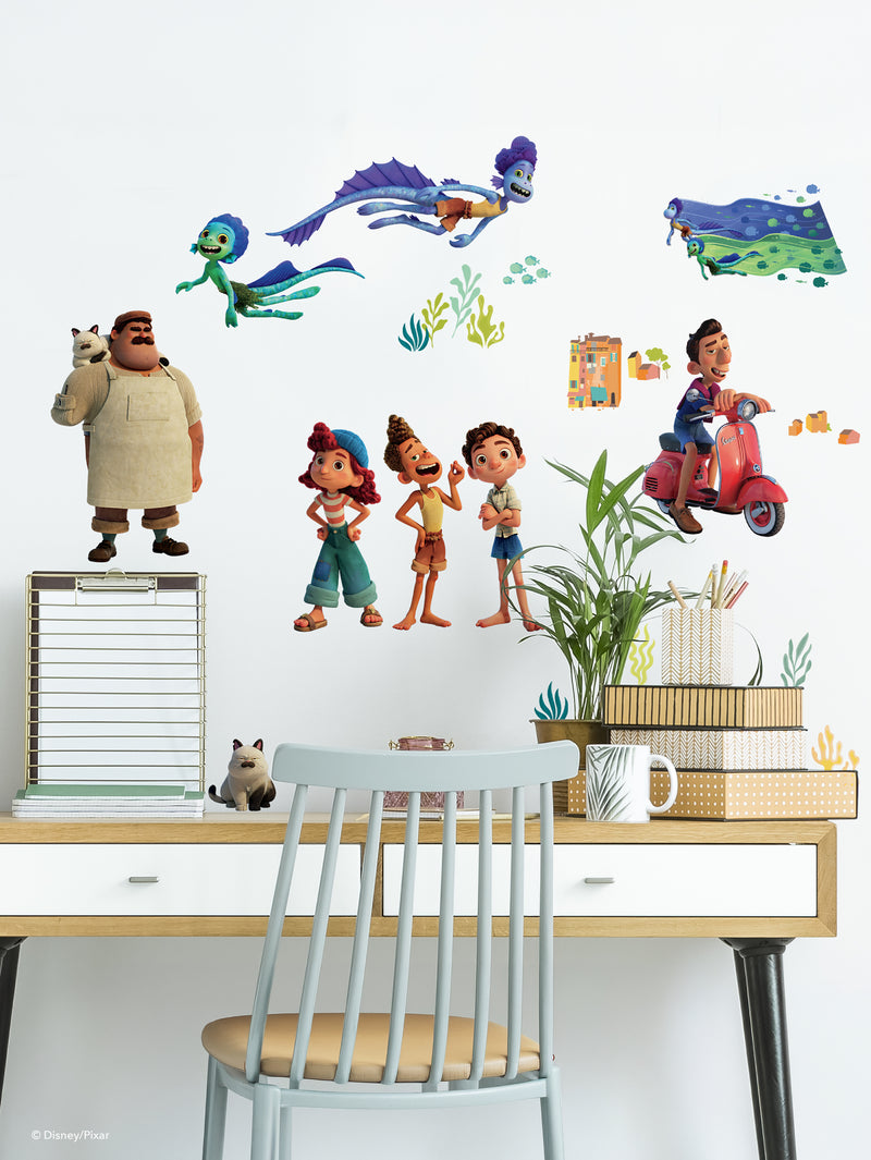 A workspace with a grey chair, white desk, stacked books, baskets, and a cup. The wall features colorful stickers of characters and scenes from Disney/Pixar’s Luca, including Luca, Alberto, Giulia, a Vespa, a cat, sea monsters, and ocean plants.
