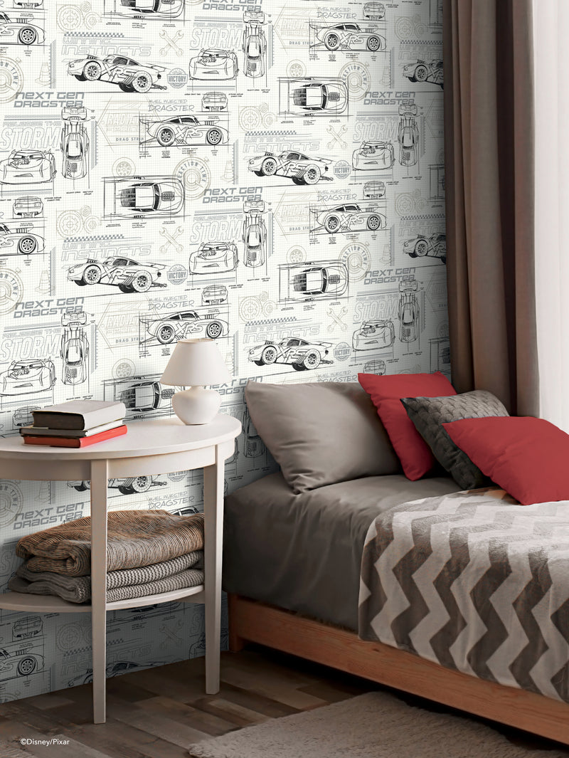 A modern bedroom with a gray and white chevron-patterned bedspread, red and gray pillows, a white round side table with books, and wall art wallpaper featuring line drawings of cars and automotive designs in black on a white background.