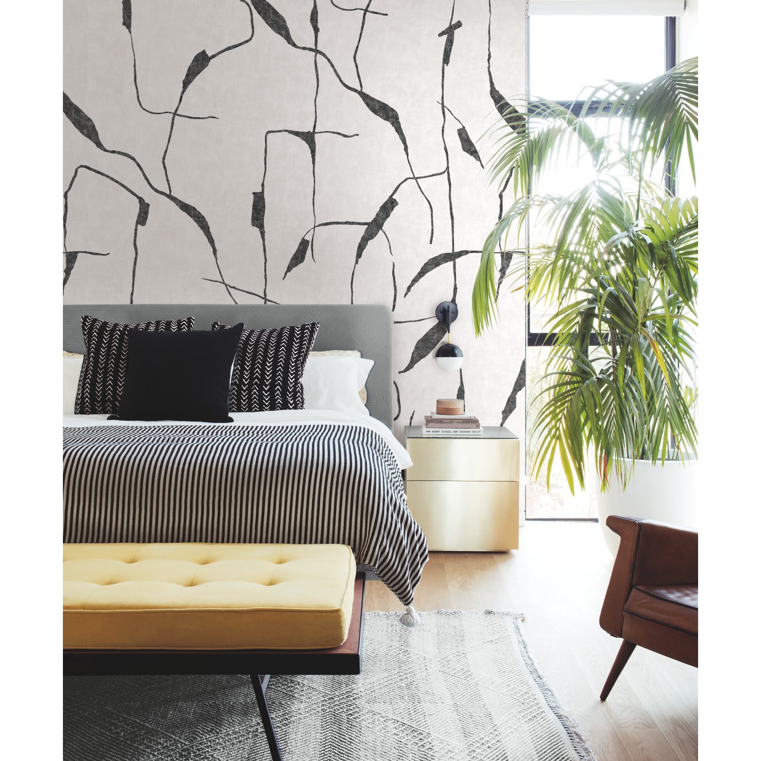 An Ivory Coast Peel & Stick Wall Mural by Nikki Chu for WallPops, featuring a beige backdrop with abstract black lines in the designer’s signature style, sets the tone; nearby, a modern velvet chair with a gold frame sits atop a gray rug beside a small stack of books topped with a dark vase of black flowers on the wooden floor.