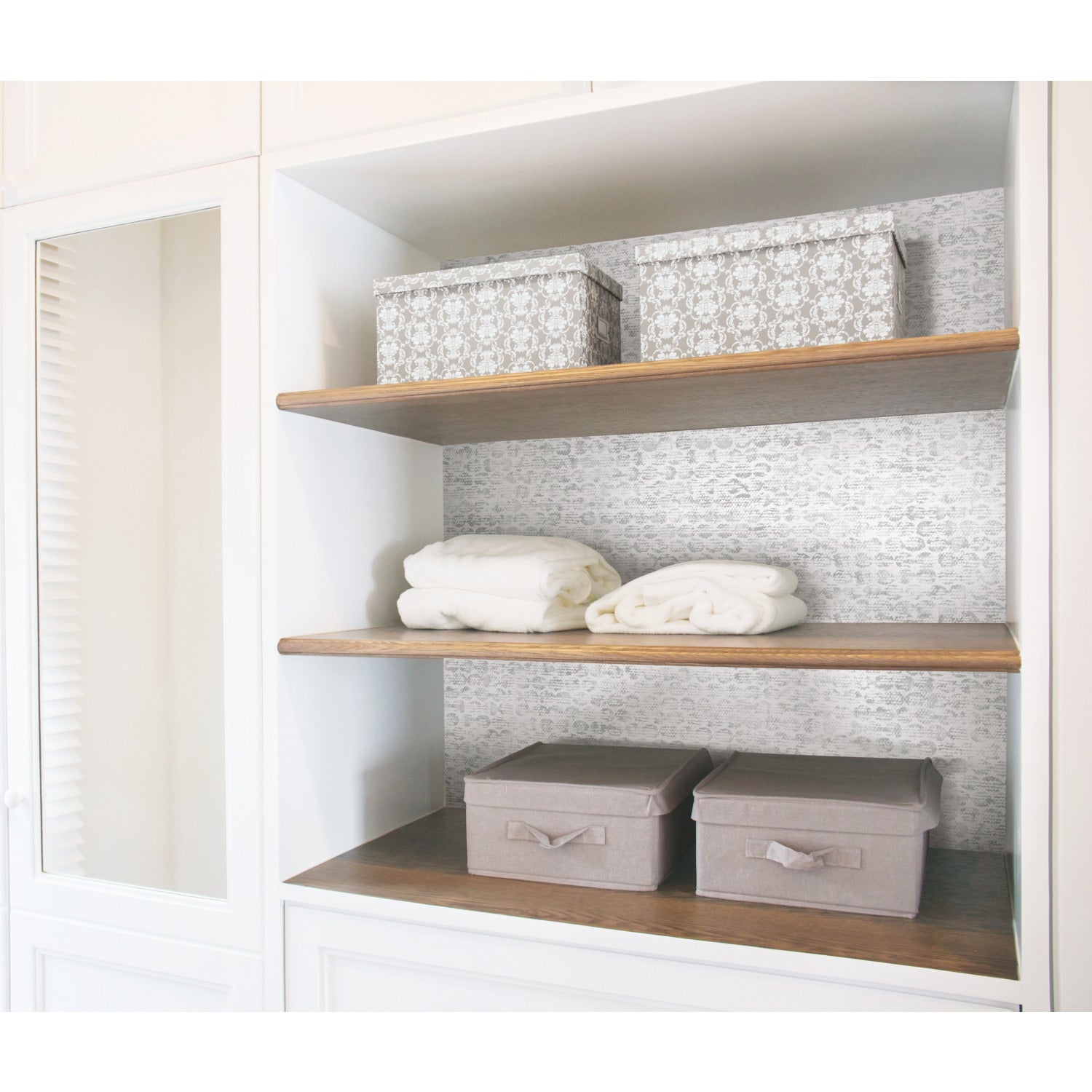 Open white shelving unit featuring two patterned boxes on the top shelf, two neatly folded white towels on the middle shelf, and two gray storage boxes with lids and handles on the bottom shelf. The shelves have a wood finish and are accented with WallPops Nikki Chu Ulo Texture Peel and Stick Wallpaper in light gray as a stylish, textured backdrop.