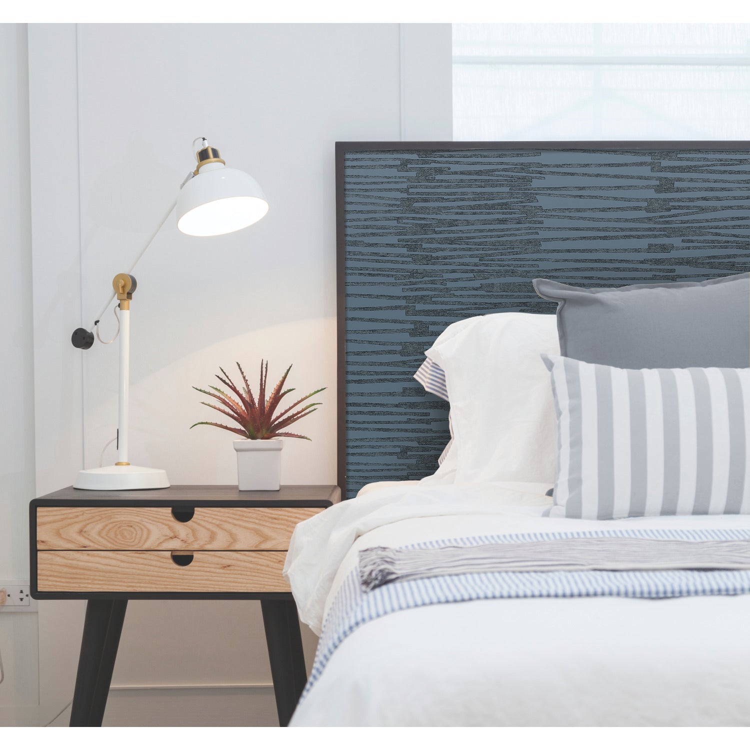 A minimal bedroom features a bed with white and gray bedding, a side table with a drawer, a potted succulent, and a white adjustable desk lamp. The headboard is blue with a textured pattern, complemented by WallPops Nikki Chu Burundi Thatch Peel and Stick Wallpaper that enhances the room’s modern appeal. The space maintains a clean look with plenty of natural light.