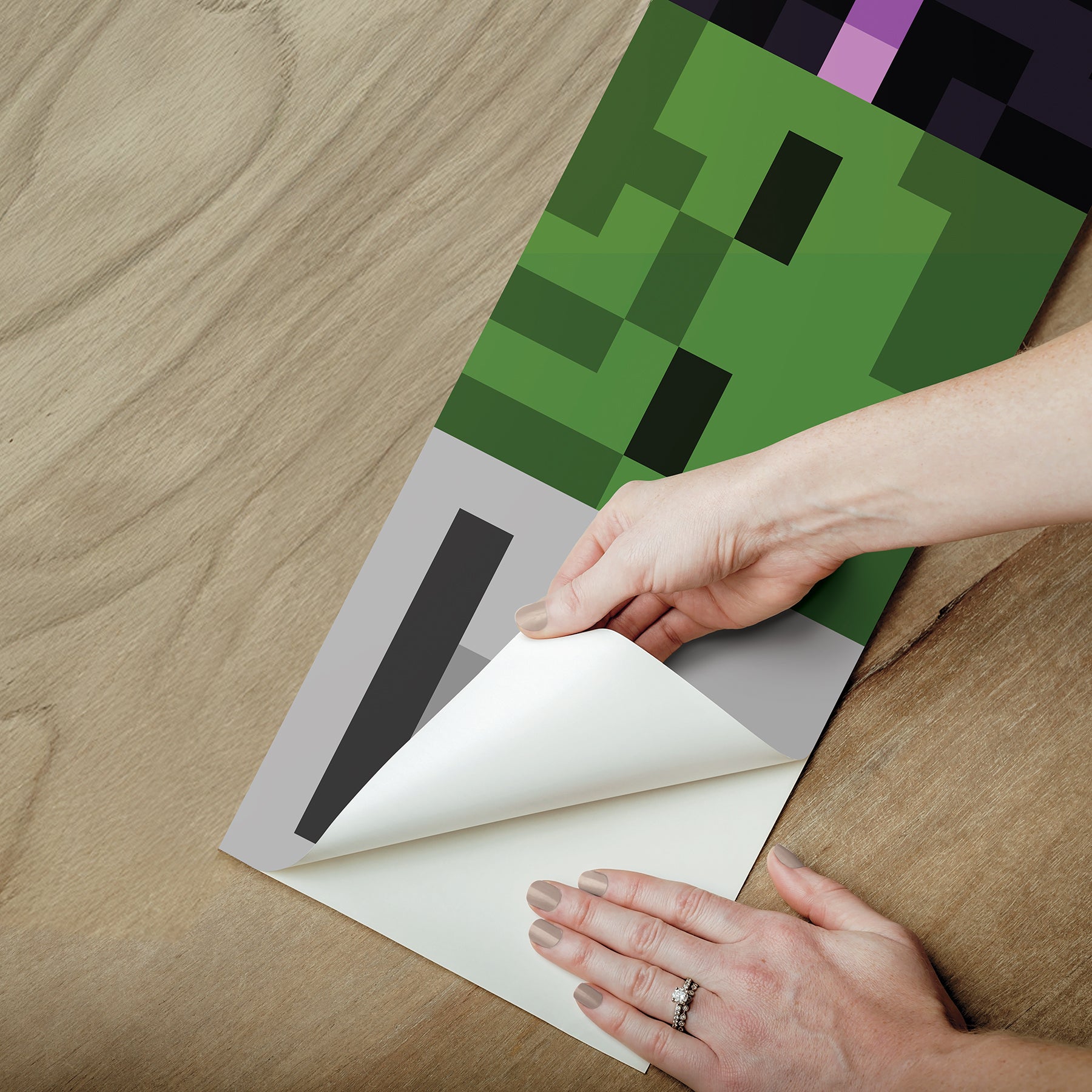 A person peels back the corner of a peel and stick Minecraft Hostile Mobs Wall Decal with a pixelated green and black design on a wooden surface.