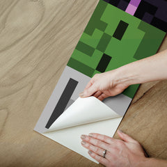 A person peels back the corner of a peel and stick Minecraft Hostile Mobs Wall Decal with a pixelated green and black design on a wooden surface.