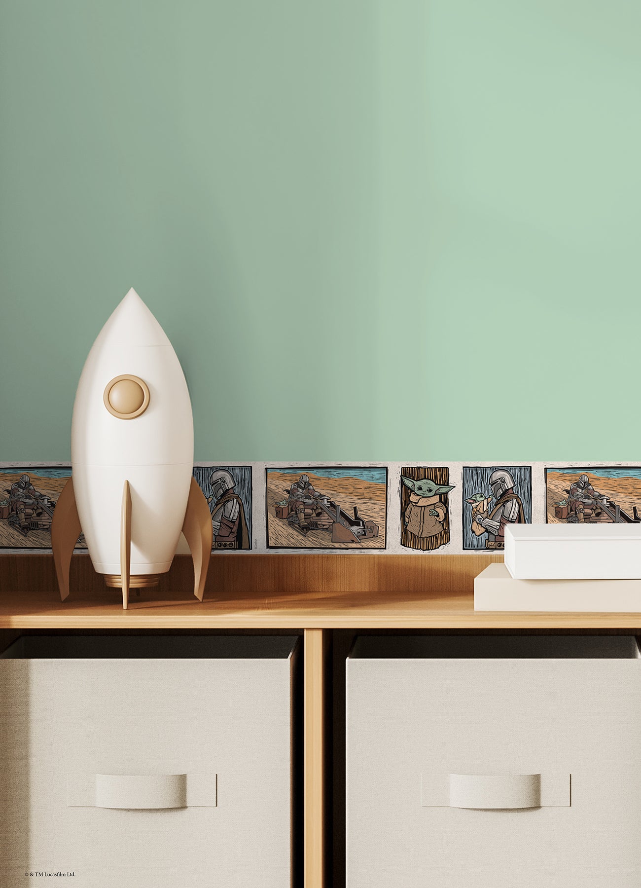 A white decorative rocket stands on a wooden shelf beside white books. Below are two beige storage bins. Behind, a light blue wall features a border of whimsical Star Wars The Mandalorian and Grogu linocut illustrations created with WallPops peel and stick wall decals. The scene is neatly arranged and playful.