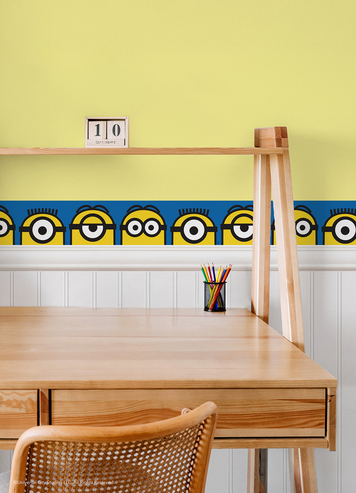 A person wearing a silver ring on their hand peels the backing off a large sticker resembling the WallPops Universal Studios Minions Always Eyeconic Border. The sticker features part of a cartoon character with yellow skin, round eyes, and black goggles set against a blue and yellow background, all laid out on a wooden surface.