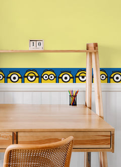 A person wearing a silver ring on their hand peels the backing off a large sticker resembling the WallPops Universal Studios Minions Always Eyeconic Border. The sticker features part of a cartoon character with yellow skin, round eyes, and black goggles set against a blue and yellow background, all laid out on a wooden surface.